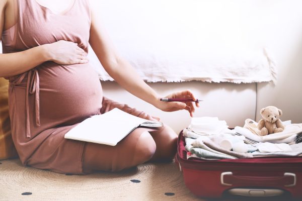 Pregnant woman packing bag for maternity hospital, making notes, checking list in diary. Expectant mother with suitcase of baby clothes and necessities preparing for newborn birth during pregnancy.